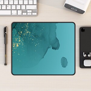 May include: A teal tablet with a black frame and a gold and teal abstract design. The tablet is on a light wood surface.