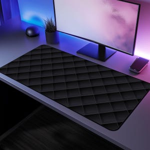 Op de afbeelding: Een zwarte gaming muismat met een ruitpatroon. De muismat ligt op een bureau met een computermonitor, een plant en een telefoon.