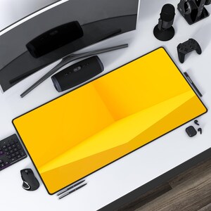 May include: A yellow desk mat with a geometric design. The mat is on a white desk with a computer monitor, a speaker, a microphone, a controller, and a pair of headphones.