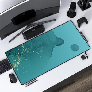 May include: A teal and gold desk mat with a watercolor-like design. The mat is on a white desk with a computer monitor, a speaker, a microphone, a game controller, and a pair of headphones.