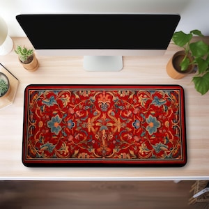 May include: A red, black and gold patterned desk mat with a floral design. The mat is on a wooden desk with a computer monitor and a potted plant.