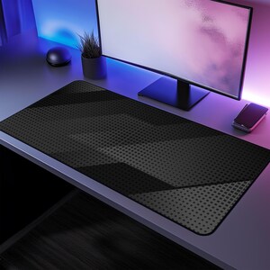 May include: A large, rectangular black mouse mat with a geometric pattern of dark grey shapes and a dotted texture. The mat is on a desk with a computer monitor, a small potted plant, and a smartphone.