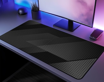 Puede incluir: Una alfombrilla de ratón negra rectangular grande con un patrón geométrico de formas gris oscuro y una textura punteada. La alfombrilla está sobre un escritorio con un monitor de ordenador, una pequeña planta en maceta y un smartphone.