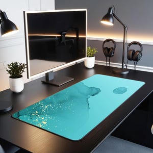 May include: A teal blue and gold desk mat with a watercolor design. The mat is on a black desk with a computer monitor, two lamps, and headphones.