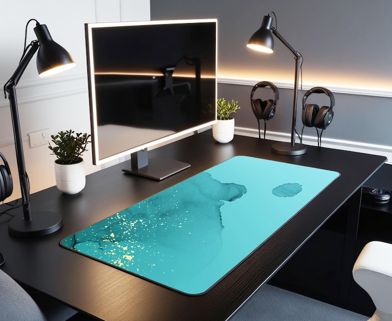 May include: A teal blue and gold desk mat with a watercolor design. The mat is on a black desk with a computer monitor, two lamps, and headphones.