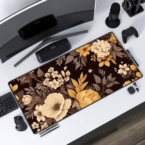 Puede incluir: Una alfombrilla de ratón grande de color negro con un diseño floral que presenta flores beige y marrones sobre un fondo negro. La alfombrilla de ratón está sobre un escritorio blanco con un monitor de ordenador, un altavoz, un mando de juego y otros accesorios.