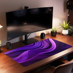 May include: A purple and black abstract design mousepad with a curved design. The mousepad is on a wooden desk with a computer monitor, a lamp, and plants.
