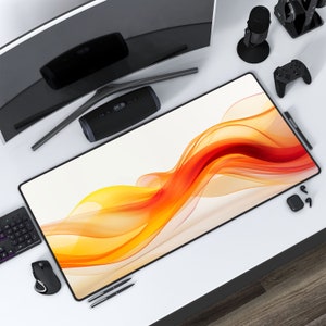 May include: A white desk mat with a colorful abstract design of orange and yellow waves. The mat is on a white desk with a black computer monitor, a black speaker, a black microphone, a black gaming controller, a black keyboard, a black mouse, and a black pen.