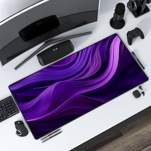 May include: Purple and blue abstract design on a large rectangular mouse pad. The mouse pad is on a white desk with a computer monitor, a speaker, a microphone, a controller, and pens.