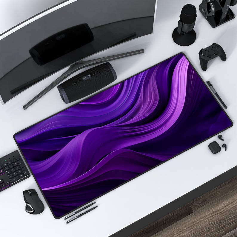 May include: Purple and blue abstract design on a large rectangular mouse pad. The mouse pad is on a white desk with a computer monitor, a speaker, a microphone, a controller, and pens.