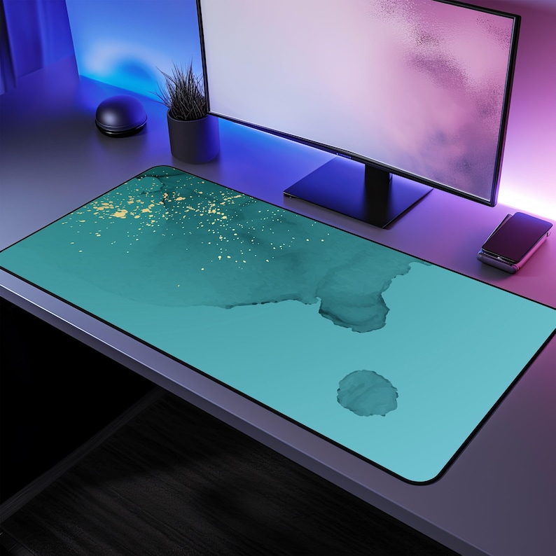 May include: A teal and gold abstract design desk mat with a smooth surface. The mat is on a desk with a computer monitor and a plant.