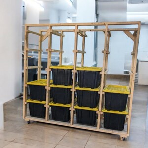 May include: A wooden storage rack on wheels, holding several black plastic bins with yellow lids. The rack has a simple, open design with multiple shelves, ideal for organizing and storing items. The bins are evenly spaced within the wooden frame.