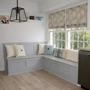 Könnte beinhalten: Eine graue Einbaubank mit mehreren dekorativen Kissen in einer Ecke eines Raumes. Die Kissen sind in Weiß-, Blau- und Beigetönen gehalten. Der Raum hat einen hellen Holzboden und ein Fenster mit gemusterten Jalousien. Eine schwarze geometrische Leuchte hängt von der Decke.