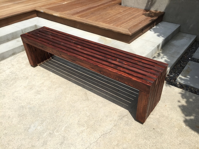 May include: A dark brown wooden bench with a slatted design. The bench is made of solid wood and has a natural finish. It is sitting on a concrete patio.