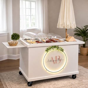 Puede incluir: Un carrito de comida blanco con una zona de servicio, tapas transparentes en forma de cúpula y una mesa auxiliar. El carrito tiene ruedas negras y un logotipo circular que dice "Artisan Bites". También se ve un paraguas decorativo y una planta pequeña.