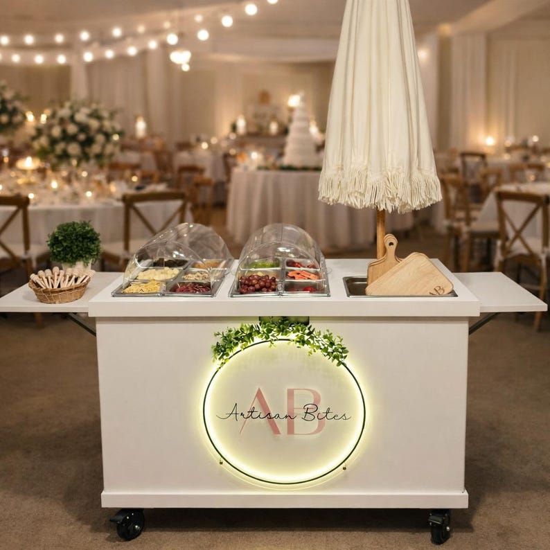May include: A white catering cart with a circular illuminated sign that reads "Artisan Bites." The cart has a white umbrella, food containers, and wooden cutting boards. The cart is on wheels and is set up in a well-lit event space.