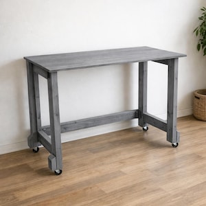 Könnte beinhalten: Ein grauer Holzschreibtisch mit rechteckiger Platte und vier Beinen. Der Schreibtisch hat ein schlichtes Design und ist zur einfachen Bewegung auf Rollen montiert. Der Schreibtisch ist aus Holz und hat eine natürliche Oberfläche.