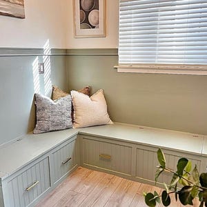 May include: A built-in bench with storage compartments and a cushion. The bench is painted a light green color and has a white trim. The bench is in a room with a window and a picture on the wall.