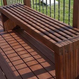 May include: A dark brown wooden bench with a slatted top and a solid base. The bench is made of stacked planks of wood and is sitting on a wooden deck.