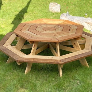 Puede incluir: Mesa de picnic octogonal de madera con una gran mesa octogonal y bancos alrededor. La mesa está hecha de madera natural con un acabado marrón claro. La mesa está situada en un césped.