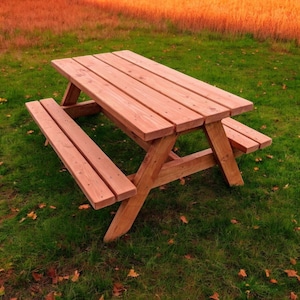 Peut inclure: Une table de pique-nique en bois avec deux bancs, en bois naturel de couleur brun rougeâtre. La table est installée sur une pelouse herbeuse avec des feuilles mortes.