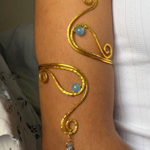 May include: Gold-toned arm cuff with a hammered texture and swirling design. The cuff features two light blue beads and a clear teardrop-shaped charm. The jewelry is worn on the arm.