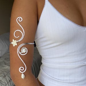 May include: Silver arm cuff with a floral and seashell design. The cuff features a delicate, swirling pattern with a small flower, a seashell, and a dangling pearl. The jewelry is worn on the upper arm.