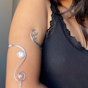 May include: A silver arm cuff with a swirling design, featuring two pearlescent beads and a dangling crystal. The jewelry is worn on the upper arm, showcasing an elegant and artistic accessory.