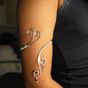 May include: A silver arm cuff with a delicate, swirling design. The cuff features two heart-shaped swirls and a curved band adorned with small, clear stones. Small blue beads dangle from the lower portion of the cuff, adding a touch of colour.