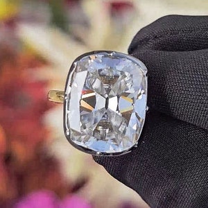 Könnte beinhalten: Ein großer, klarer, kissenförmiger Diamantring, der in eine goldene Fassung gefasst ist. Der Ring befindet sich auf einem schwarzen Stoffhintergrund.