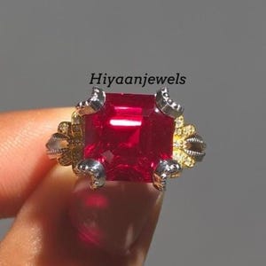 Könnte beinhalten: Nahaufnahme eines quadratisch geschliffenen, leuchtend roten Edelsteinrings. Der Ring weist aufwendige Silber- und Golddetails um den Edelstein auf. Der Text "Hiyaanjewels" ist sichtbar.