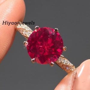 Pode incluir: Um anel em ouro rosa com uma grande pedra preciosa redonda e vermelha escura. A banda é adornada com pequenas pedras preciosas transparentes. O texto "Hiyaanjewels" é visível no anel.