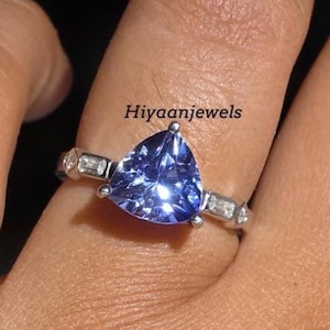Op de afbeelding: Een zilveren ring met een grote, driehoekige, blauwe edelsteen. De ring is gezet met kleinere, rechthoekige, heldere stenen. De tekst "Hiyaanjewels" is zichtbaar op de afbeelding.
