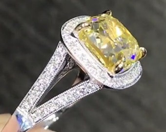 May include: A yellow cushion-cut diamond engagement ring with a halo setting and a split shank. The ring is set in white gold and features a row of smaller diamonds on the band.