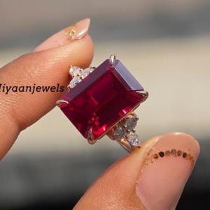 Könnte beinhalten: Ein goldener Ring mit einem großen, rechteckigen, tiefroten Edelstein. Der Ring ist mit kleineren, klaren Edelsteinen verziert. Der Text "Hiyaanjewels" ist auf dem Ring zu sehen.