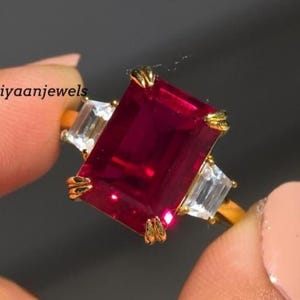 Könnte beinhalten: Ein goldener Ring mit einem großen, rechteckigen, tiefroten Edelstein, flankiert von zwei kleineren, rechteckigen, klaren Edelsteinen. Der Ring wird von den Fingern einer Person gehalten. Der Text "Hiyaanjewels" ist sichtbar.