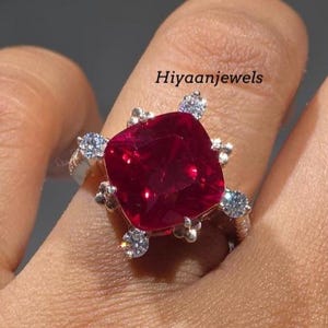 Könnte beinhalten: Ein silberner Ring mit einem großen, quadratisch geschliffenen, tiefroten Edelstein im Mittelpunkt. Der Ring ist mit kleineren, klaren Edelsteinen um den zentralen Stein verziert. Der Text "Hiyaanjewels" ist sichtbar.