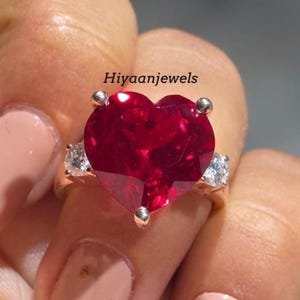 Könnte beinhalten: Ein goldener Ring mit einem großen, herzförmigen, tiefroten Edelstein. Zwei kleinere, klare Edelsteine flankieren den herzförmigen Stein. Der Ring wird von einer Hand gehalten, im Hintergrund steht der Text "Hiyaanjewels".
