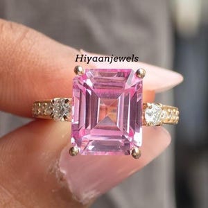 Könnte beinhalten: Ein goldener Ring mit einem großen, rechteckigen rosa Edelstein. Das Band ist mit kleineren, klaren Edelsteinen verziert. Der Ring wird hochgehalten, um sein Design und seinen Glanz zu zeigen.
