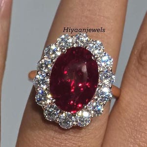 Può includere: Un anello in oro rosa con un grande rubino ovale circondato da pietre preziose chiare più piccole. L'anello è su un dito, mostrando il design e la brillantezza. Il testo "Hiyaanjewels" è visibile.