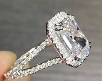May include: A silver engagement ring with a large, square-shaped diamond set in a halo setting. The ring has a split shank with pave diamonds.