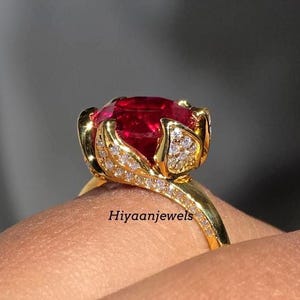 Könnte beinhalten: Ein goldener Ring mit einem großen, oval geschliffenen, leuchtend roten Edelstein. Die Fassung ist mit kleinen, funkelnden weißen Steinen und einem floralen Design verziert. Der Ring ist aus Gold. Der Text "Hiyaanjewels" ist sichtbar.