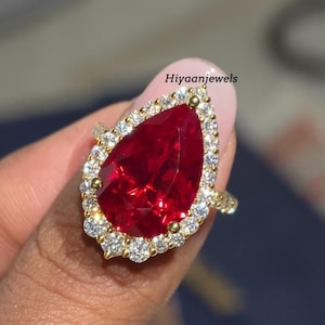 Könnte beinhalten: Ein goldener Ring mit einem großen, birnenförmigen, tiefroten Edelstein, umgeben von kleineren, klaren Edelsteinen. Der Ring wird vor einem rosa Hintergrund gehalten. Der Text "Hiyaanjewels" ist sichtbar.