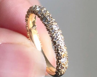 Può includere: Un anello d'oro con una fascia interamente incastonata di piccole pietre preziose trasparenti. L'anello è tenuto in mano e le pietre preziose brillano alla luce. È un gioiello delicato.