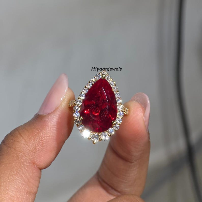 Pu&ograve; includere: Un anello d'oro con una grande pietra rossa a forma di pera, circondata da un alone di pietre pi&ugrave; piccole e chiare. L'anello &egrave; tenuto in mano. Il testo "Hiyaanjewels" &egrave; visibile.