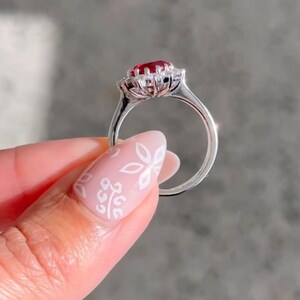 May include: A silver ring featuring a red gemstone encircled by smaller white gemstones. The ring is set in a floral design.