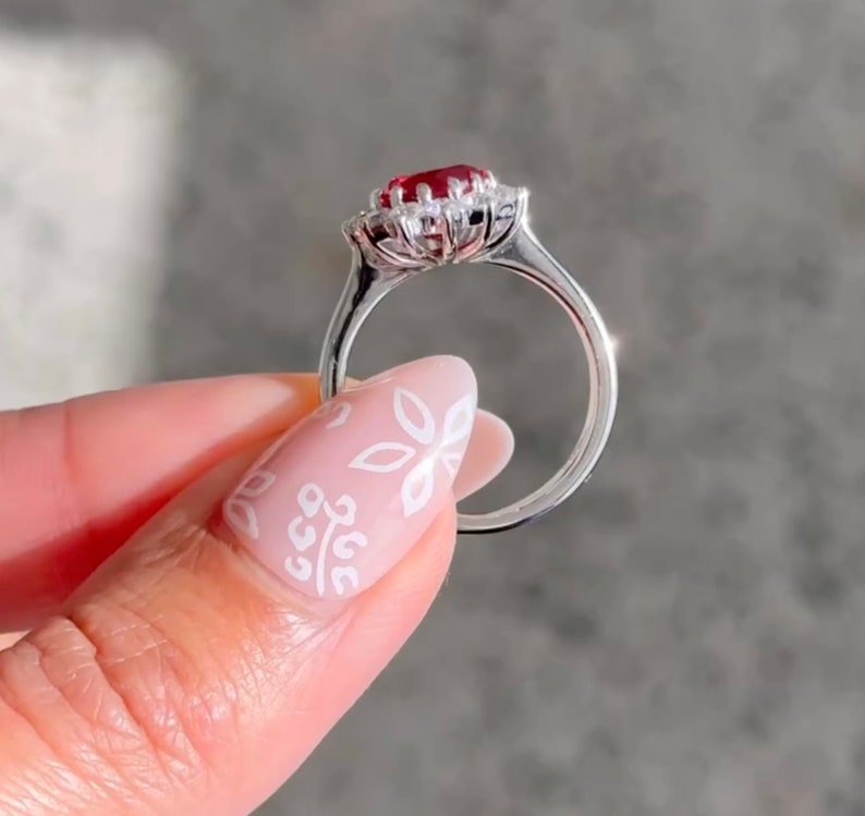 May include: A silver ring featuring a red gemstone encircled by smaller white gemstones. The ring is set in a floral design.