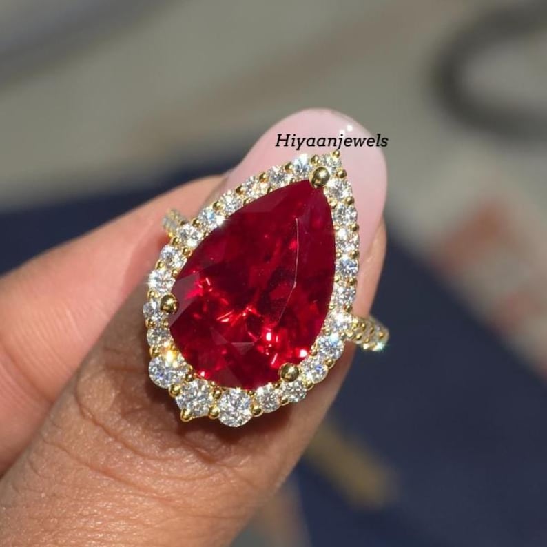 Pu&ograve; includere: Un anello d'oro con una grande pietra preziosa rossa a forma di pera, circondata da pietre preziose pi&ugrave; piccole e chiare. L'anello &egrave; tenuto su uno sfondo rosa. Il testo "Hiyaanjewels" &egrave; visibile.