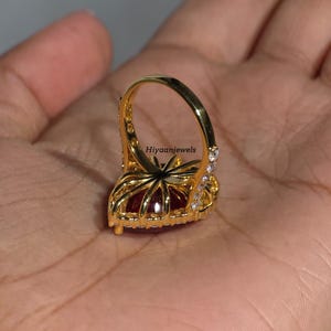 Pu&ograve; includere: Un anello color oro con una grande pietra preziosa rosso scuro a forma di cuore. L'anello &egrave; incastonato con piccole pietre trasparenti attorno alla gemma principale e lungo la fascia. Il testo "Hiyaanjewels" &egrave; visibile.