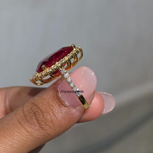 Pu&ograve; includere: Un anello d'oro con una grande pietra preziosa rosso scuro a forma di pera, circondata da un alone di pietre pi&ugrave; piccole e chiare. La fascia &egrave; anch'essa ornata da piccole pietre chiare. L'anello &egrave; tenuto in mano, mostrando il suo design.